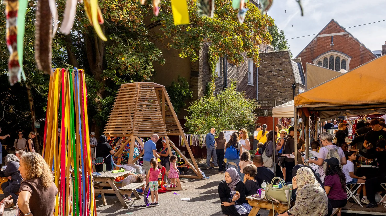 Festival crowd sitting at tables around the timber shading pavilion with colourful ribbons and market stalls