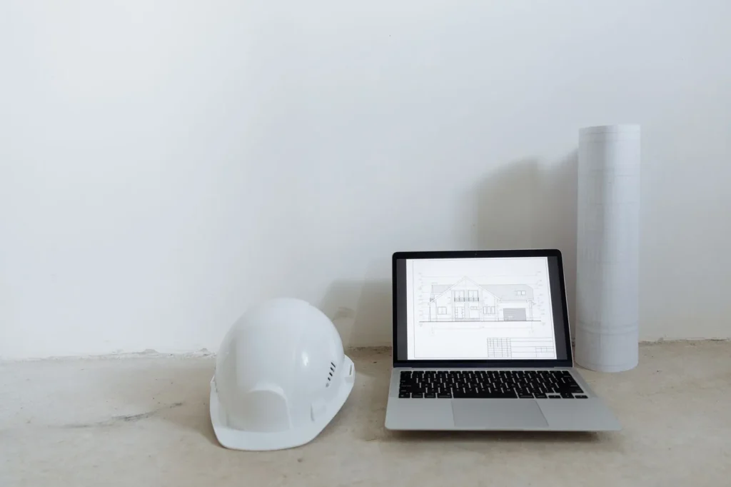 Laptop showing a building elevation drawing beside a white hard hat and a rolled drawing