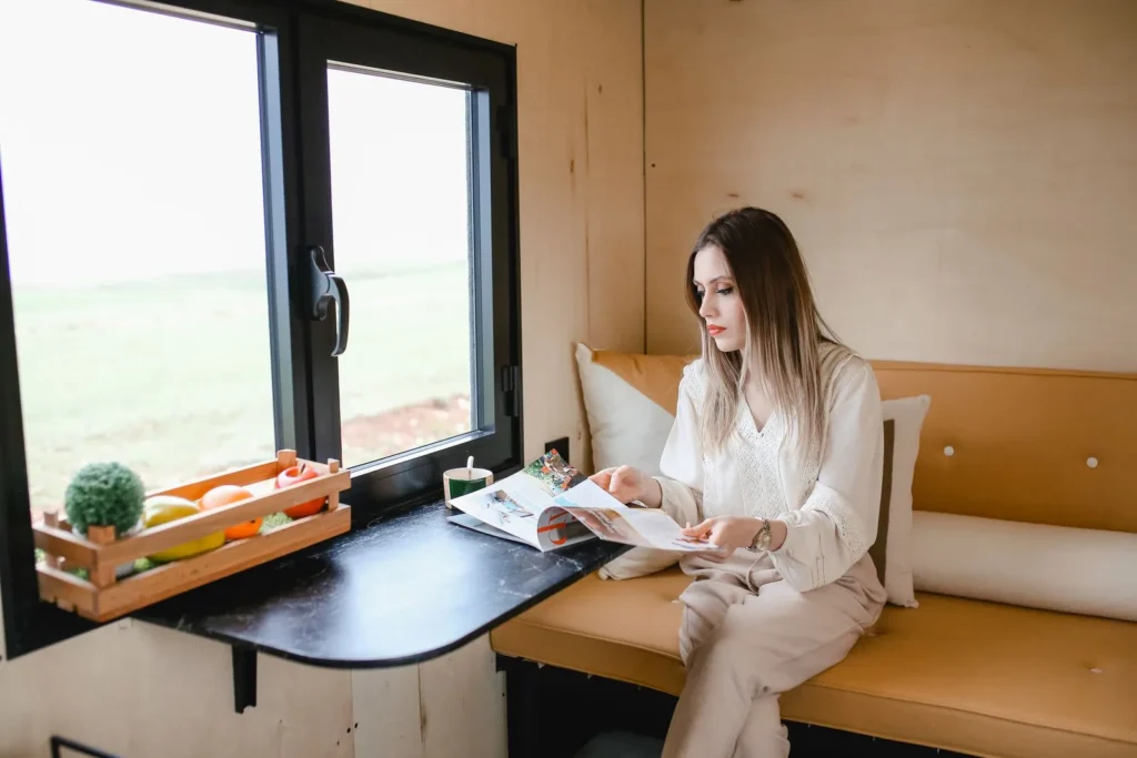 Woman reading at a fold-down table by a large window inside a plywood-lined tiny house