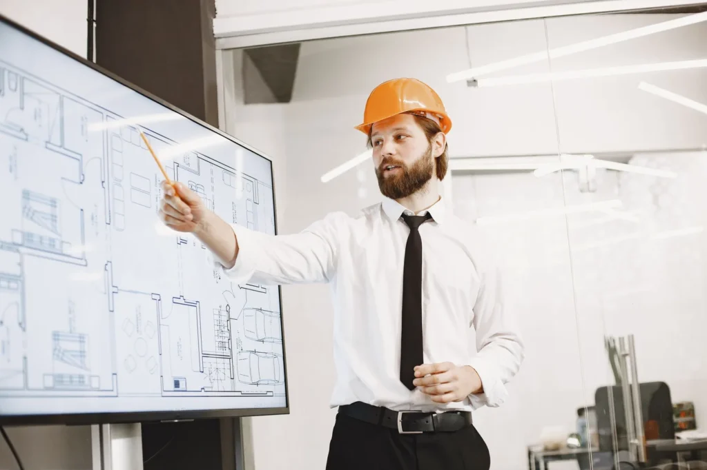 Architect in a hard hat presenting plan drawings on a large screen, where AI rendering for architecture supports client reviews