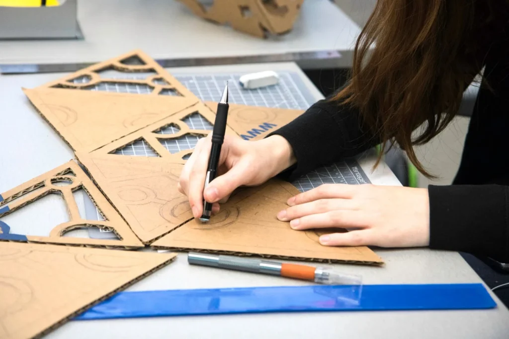 Student drawing shapes onto cardboard beside a craft knife, a ruler and cut out model parts