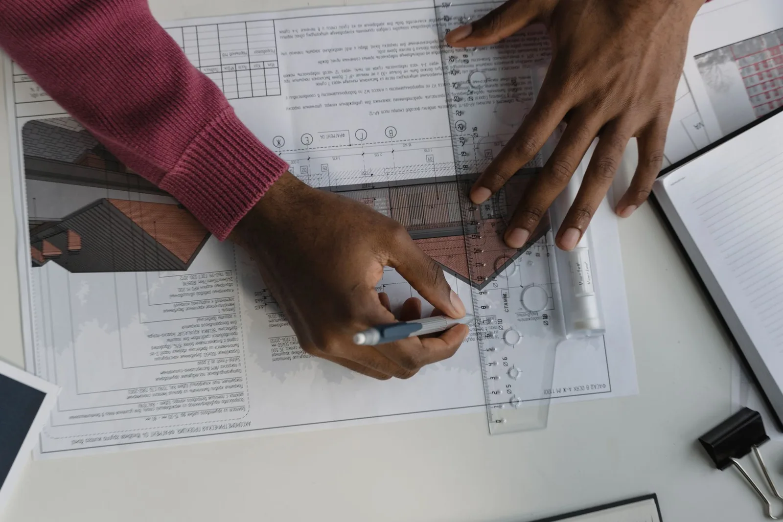 Hands drafting with a pencil and set square on a printed architectural drawing sheet with a house rendering