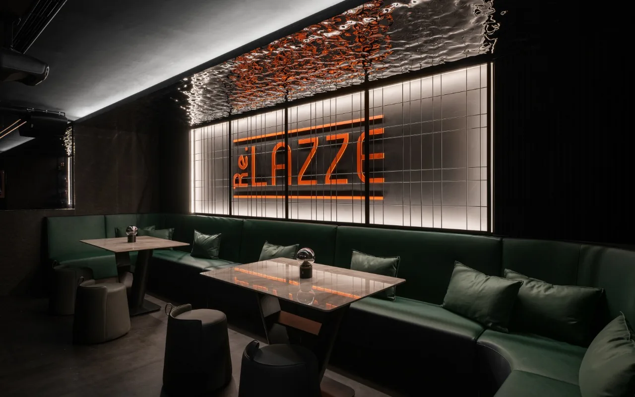 Dark bar interior with green leather banquettes, stone-topped tables and a glowing orange ReLazze sign behind a gridded glass screen