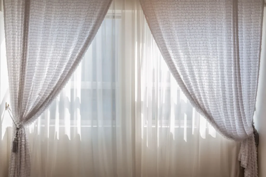 Sheer white curtains tied back from a bright window, filtering daylight into a room