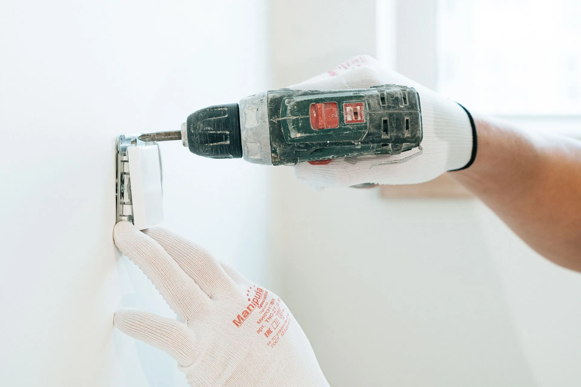 Gloved hands fixing a wall socket in place with a cordless drill on a white wall