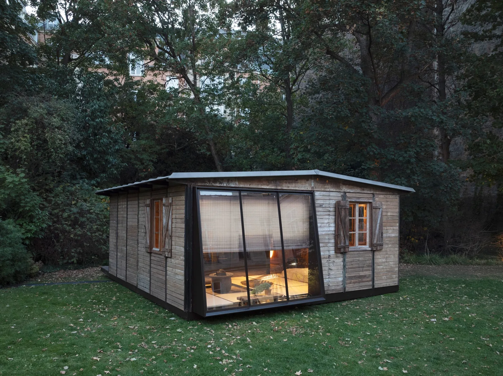 Glazed end wall of the timber house with bamboo blinds, showing a lit interior with low furniture
