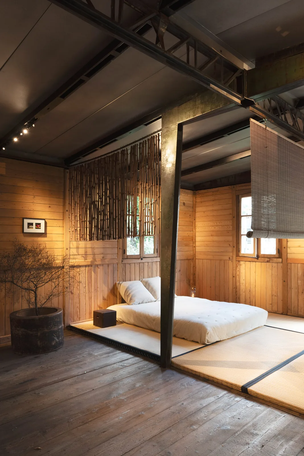 Corner of the timber interior with a hanging bamboo screen, a potted tree, a low bed and a steel roof beam