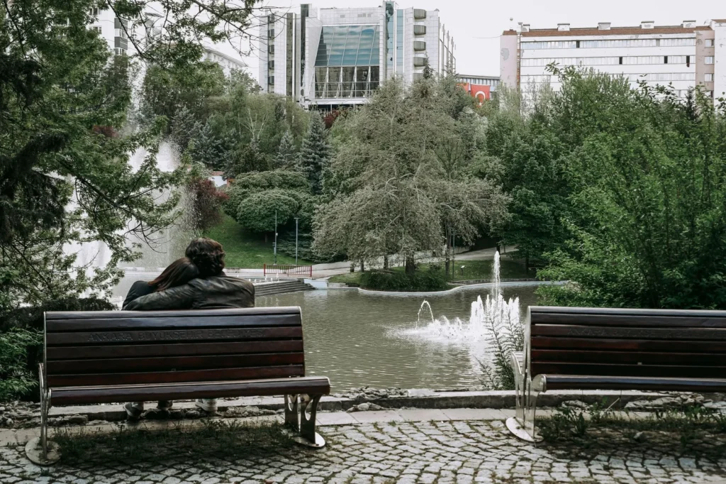 Wooden benches facing a pond with a fountain in a city park surrounded by tall buildings