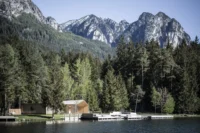 Timber lake hut and changing block on the shore below larch forest and jagged peaks, Lake House Völs by NOA