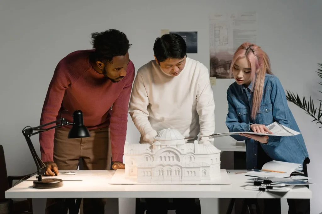 Three designers leaning over a white architectural model on a lit table while discussing the scheme
