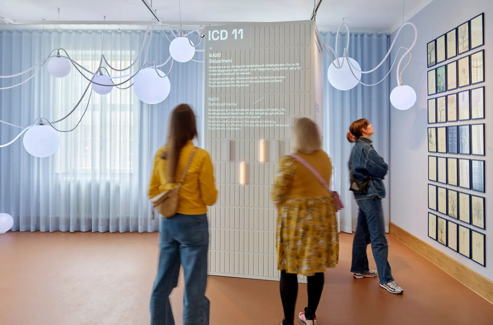 Visitors in a bright exhibition room with sheer curtains, hanging lights and wall displays