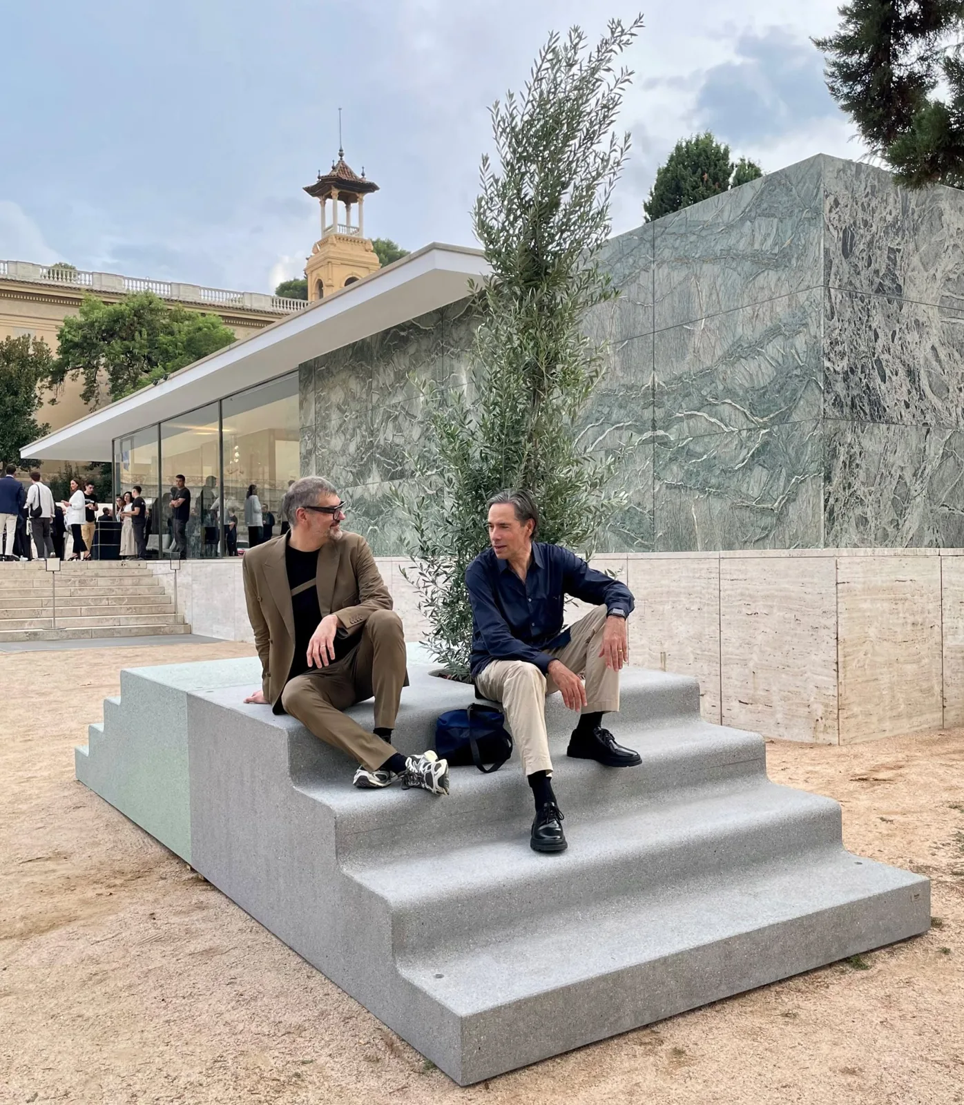 Two men sitting on pale grey concrete steps in front of the glass and marble Barcelona Pavilion