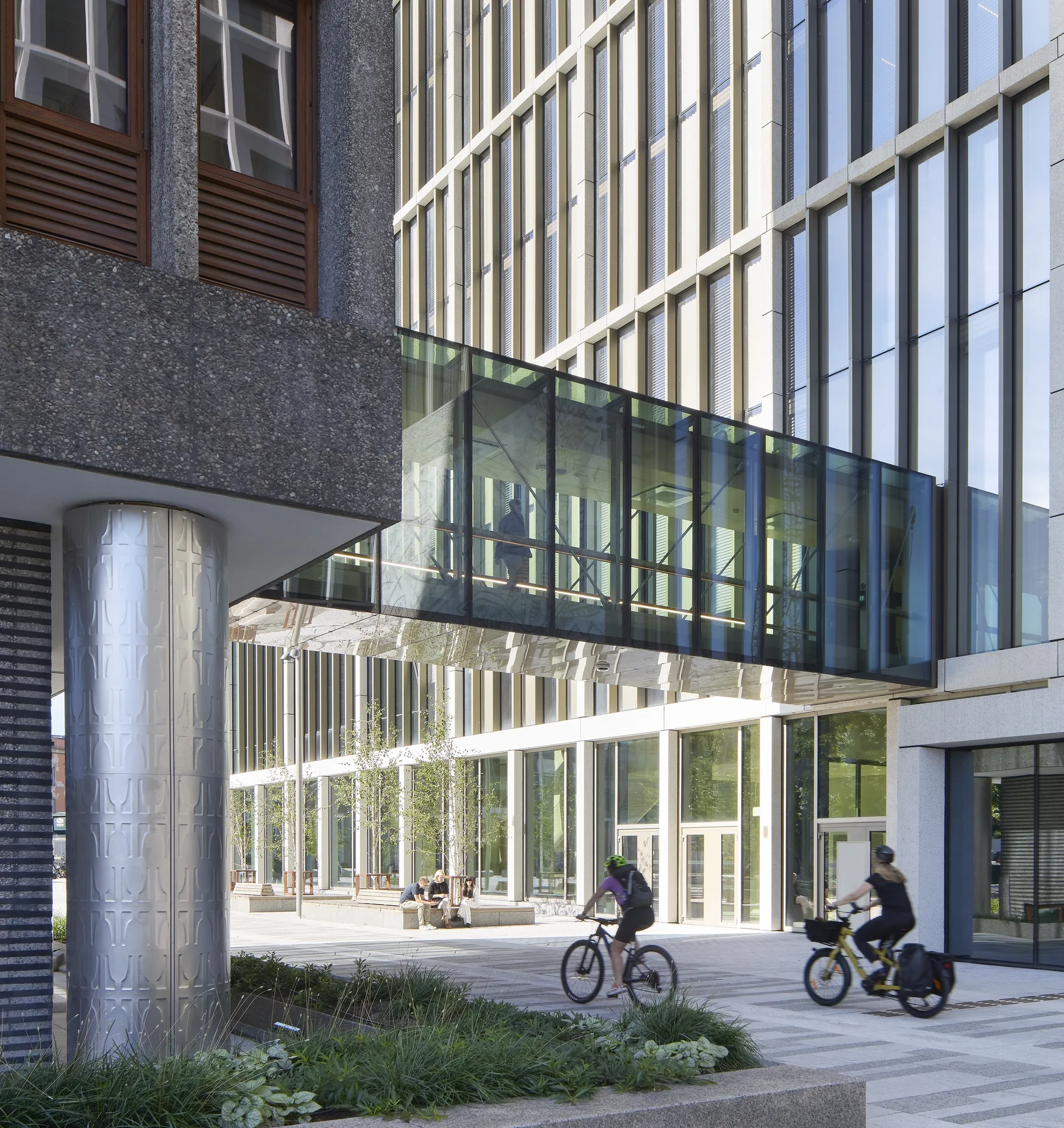 Glazed connecting bridge spanning between Høyblokken and a new block as cyclists ride across the plaza below, New Government Quarter by Nordic Office of Architecture