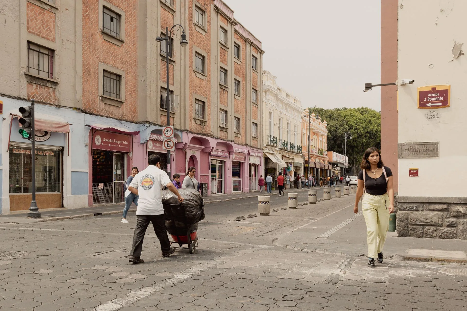 Pedestrians on a cobbled street in Puebla, Mexico, a woman walking and a man pushing a loaded cart past pink facades
