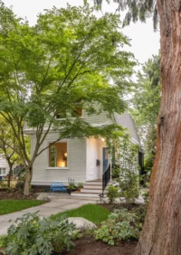 Front garden with a stone path and a tall tree trunk in front of the white clapboard cottage, Phinney Mini by Best Practice Architecture
