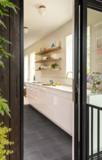 Narrow white kitchen with open oak shelves seen through the folding glass door from the deck, Phinney Mini by Best Practice Architecture