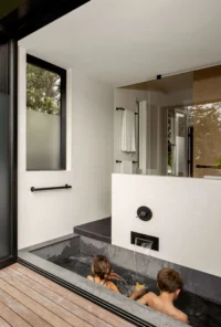 Two children in the sunken stone ofuro, opened to the timber deck through a folding glass door, Phinney Mini by Best Practice Architecture