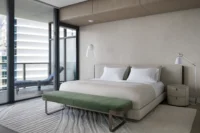Primary bedroom with a long headboard, green upholstered bench and a wavy carved rug by the balcony doors, PS Apartment by Nar Design Studio