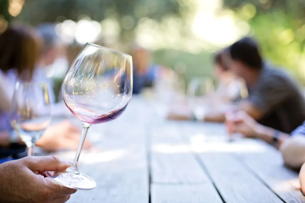 Guest holding a glass of red wine at a long wooden table set outdoors, the shared ritual behind the raised table landscape idea
