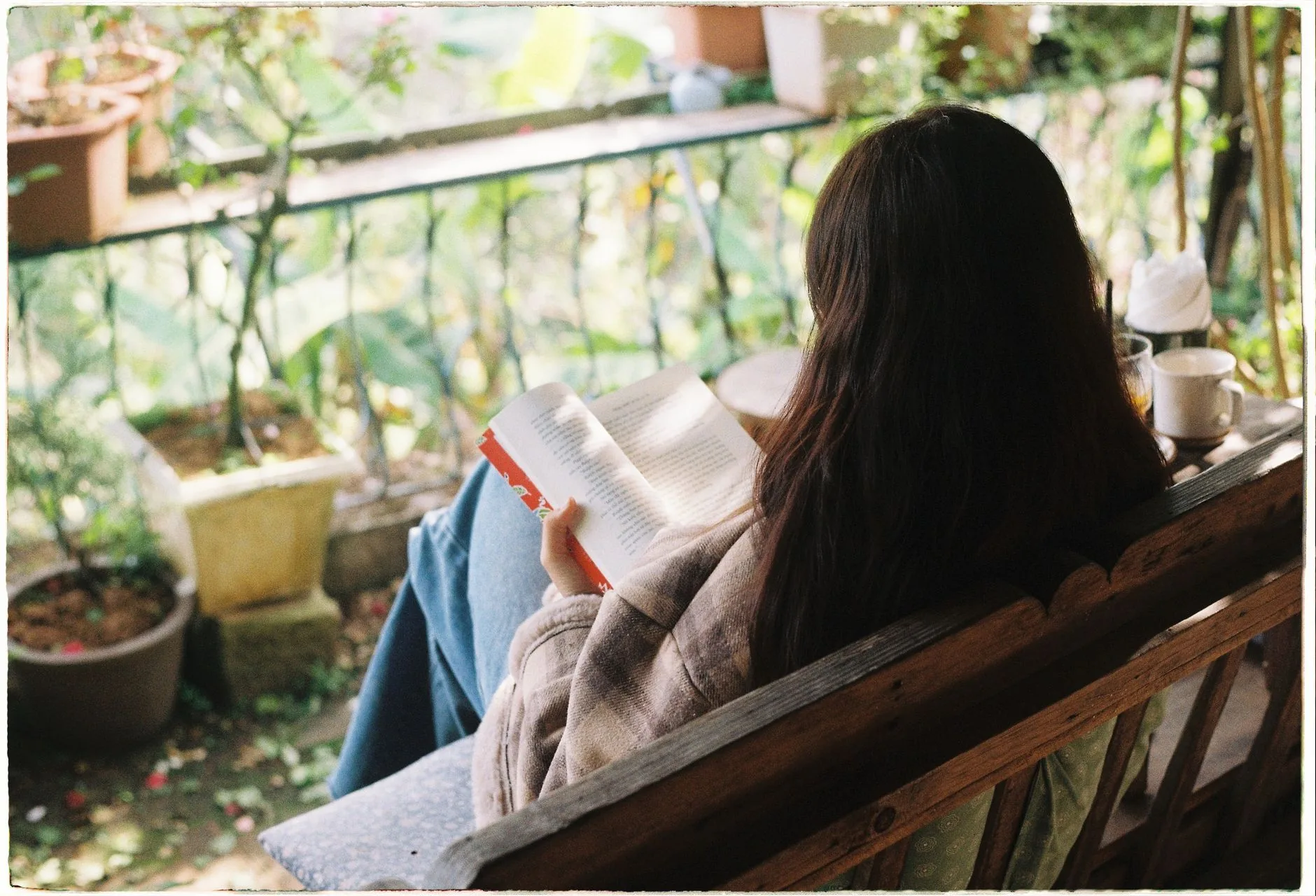 Woman reading a book on a wooden bench surrounded by potted plants on a garden terrace