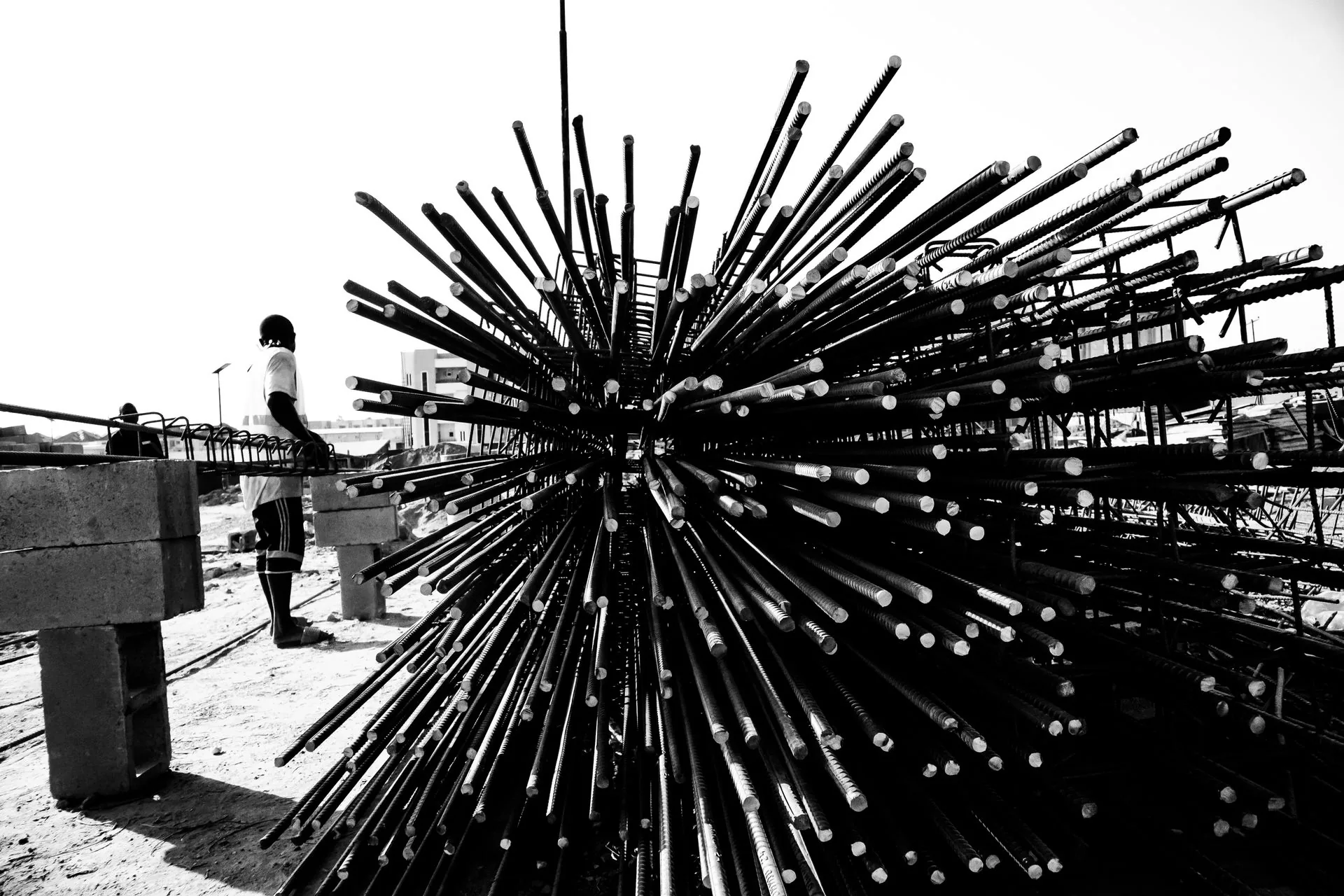 Black and white view of a large bundle of steel rebar stacked on concrete blocks with a worker on site