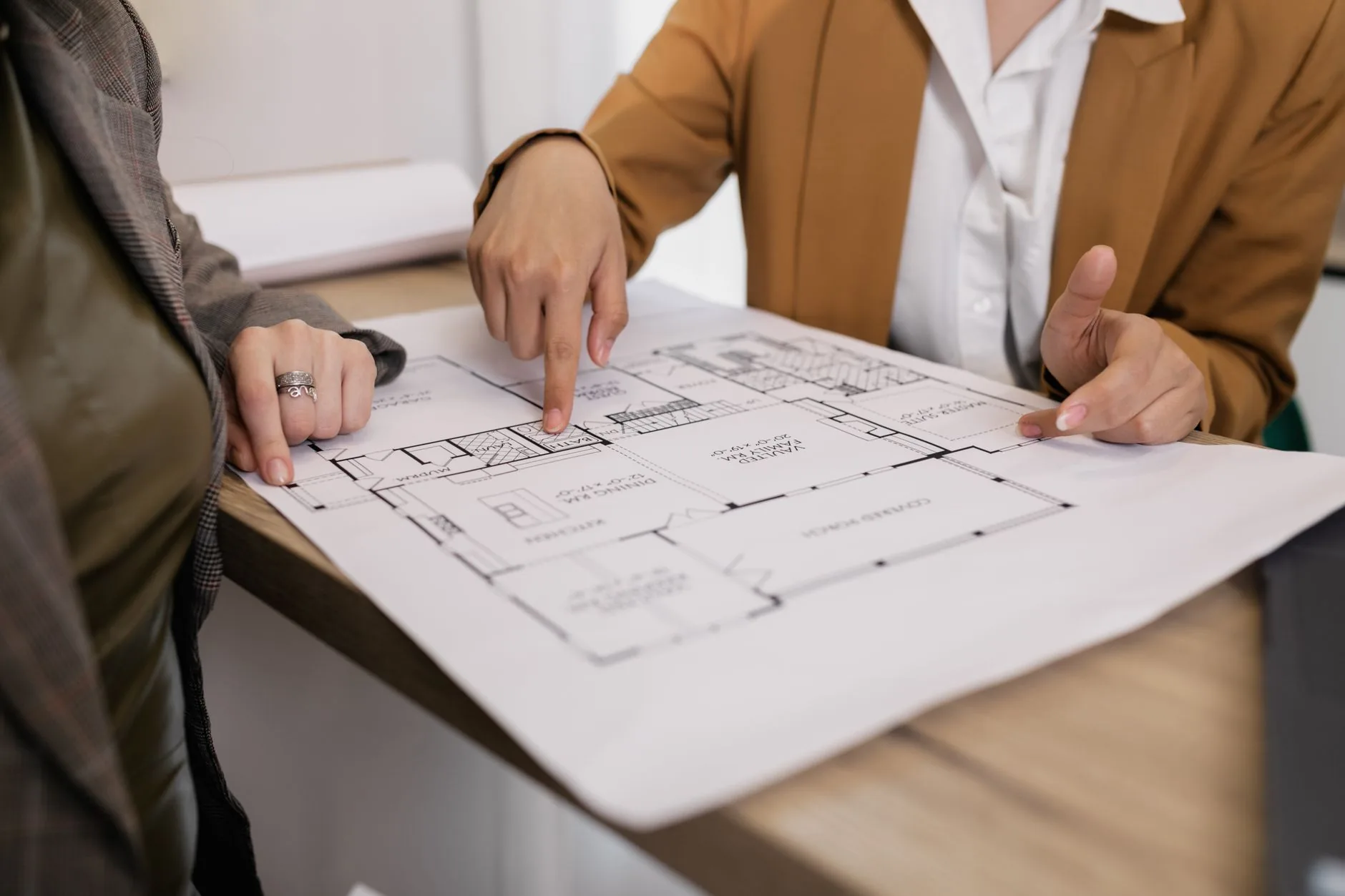 Two professionals point at a printed house floor plan with labelled rooms on a wooden table