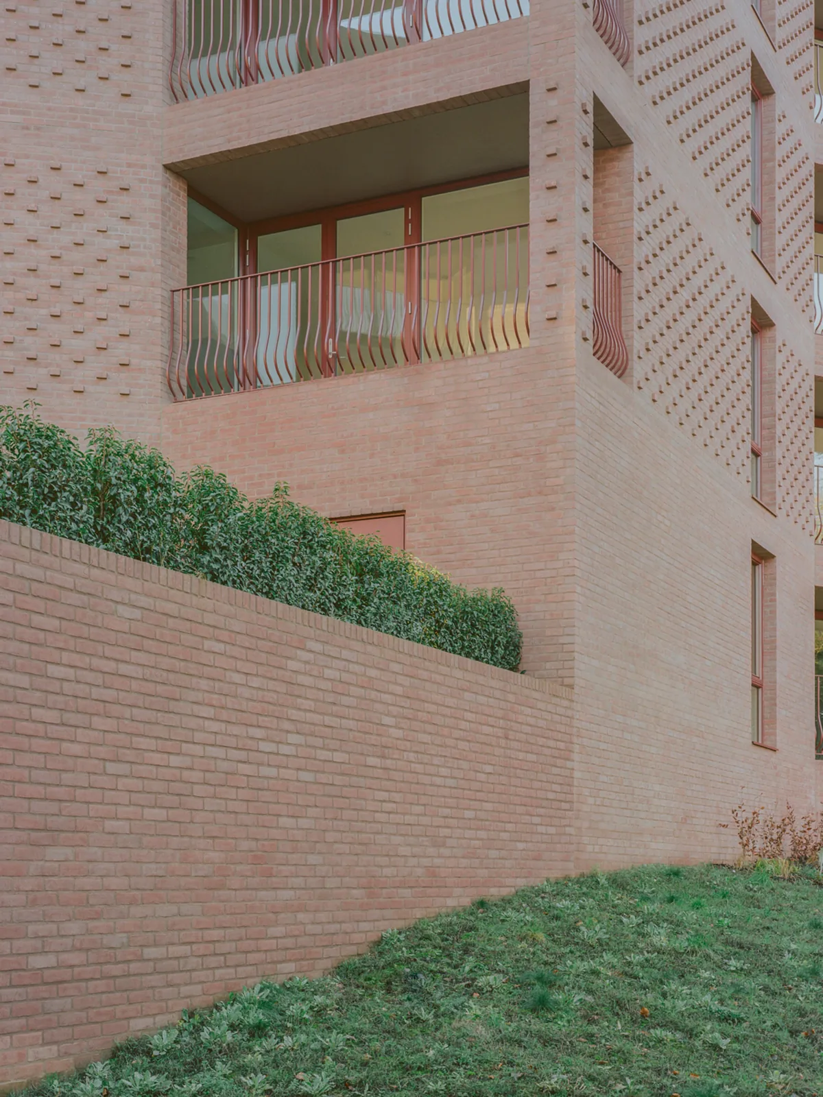 Corner of a pale brick block with a recessed balcony, perforated brickwork and a hedge along a raised terrace