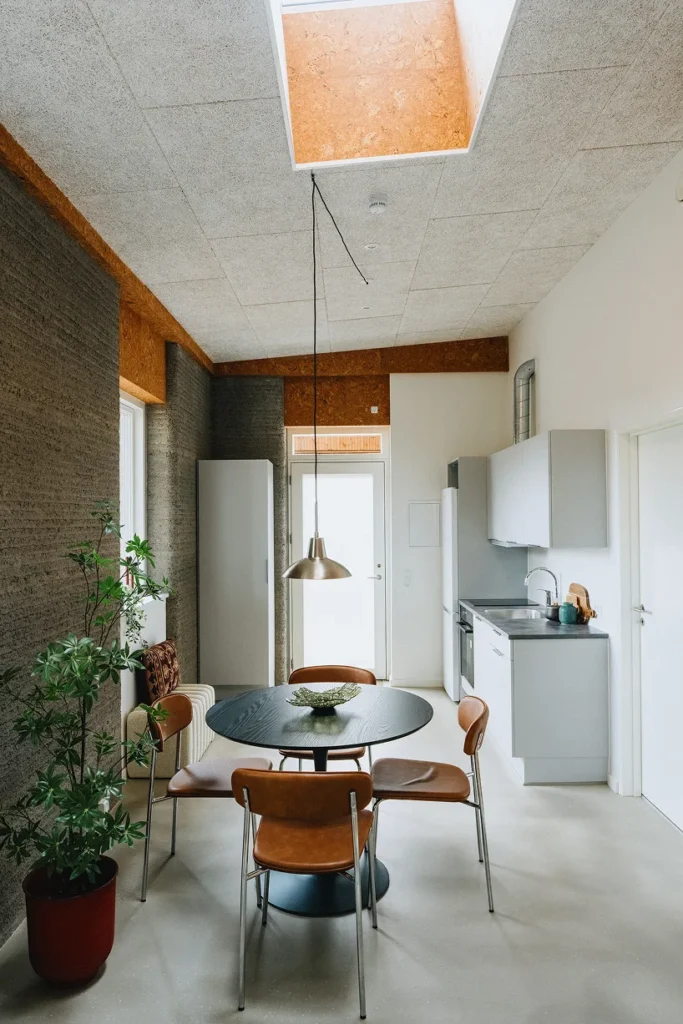 Kitchen and dining corner inside one of the 3D printed student homes, lit by a roof window above textured printed concrete walls