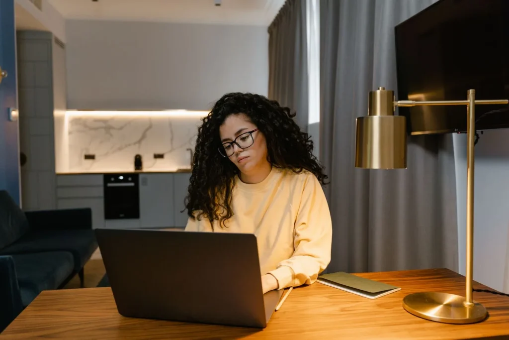 Woman working on a laptop at a wooden desk at home in the evening, one of many side hustles for architects done after hours