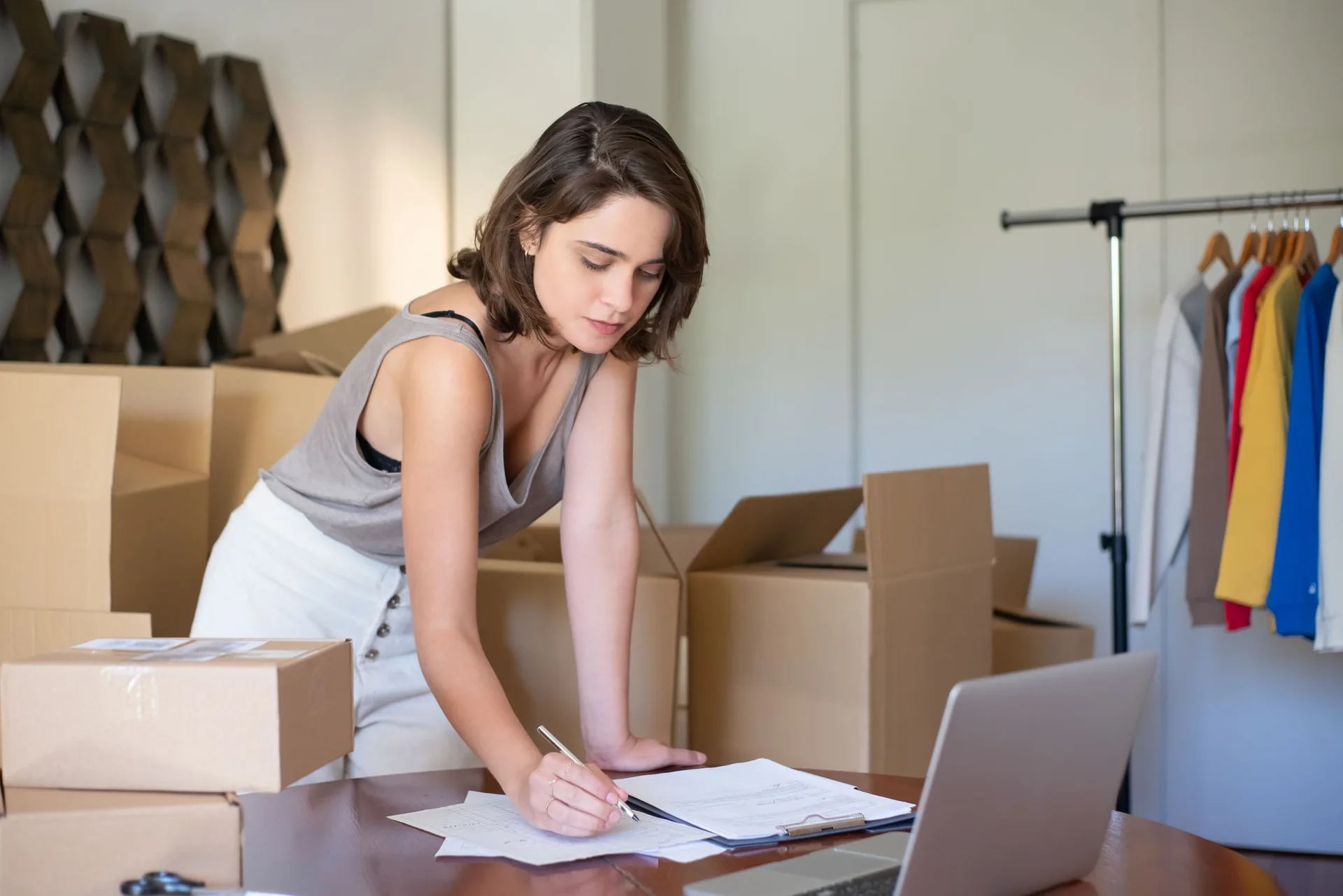 Small online clothing business owner writing on order sheets beside a laptop, shipping boxes and a clothes rack