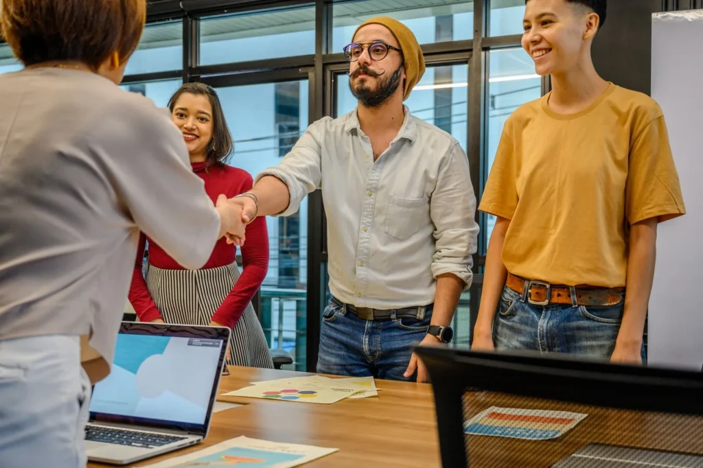 Colleagues shaking hands across a meeting table with a laptop and printed sheets