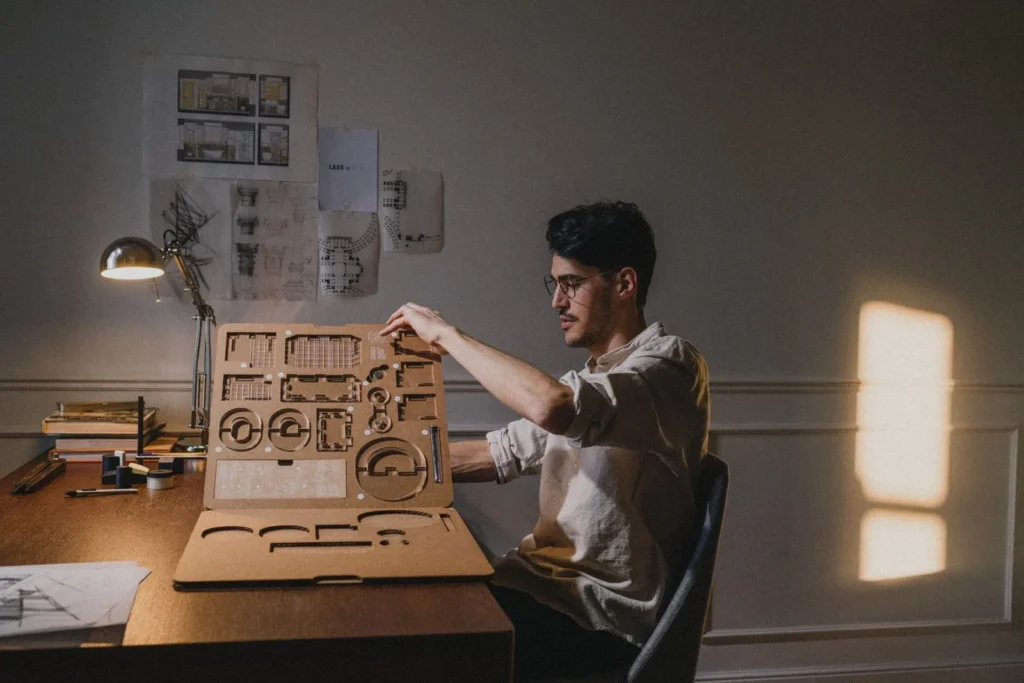 Architect assembling a laser-cut cardboard model at a desk under a lamp, with drawings pinned on the wall