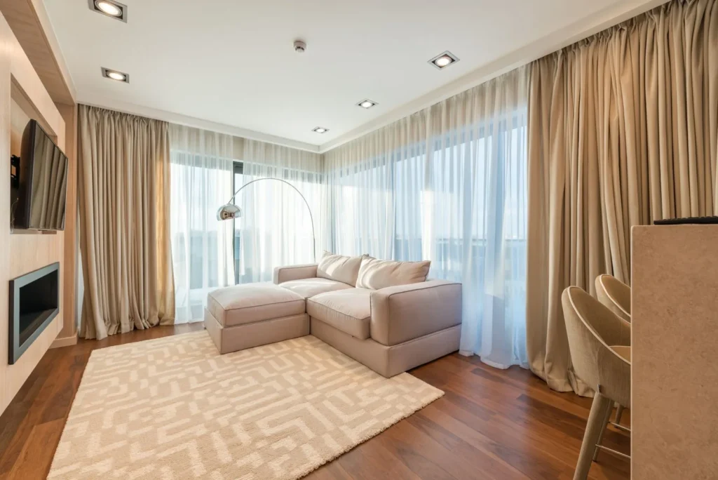 Warm minimalist living room with a cream sectional sofa, sheer curtains, a patterned rug and wood floors, reflecting American interior design trends