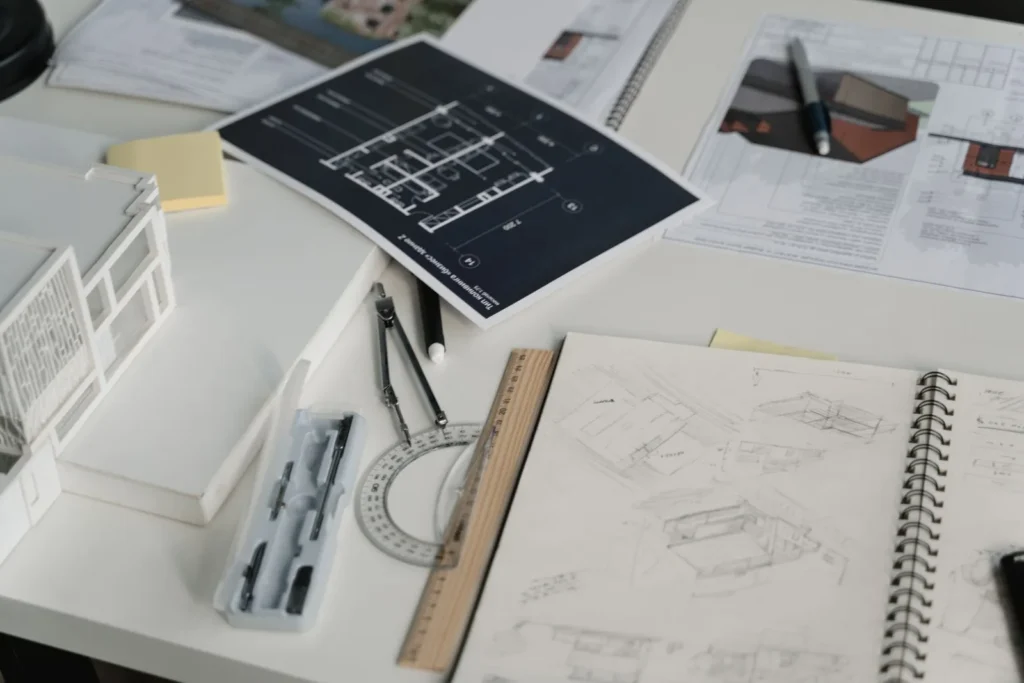 Desk covered with printed plans, a sketchbook, drafting tools and a white study model, practice work alongside 3ds Max YouTube tutorials