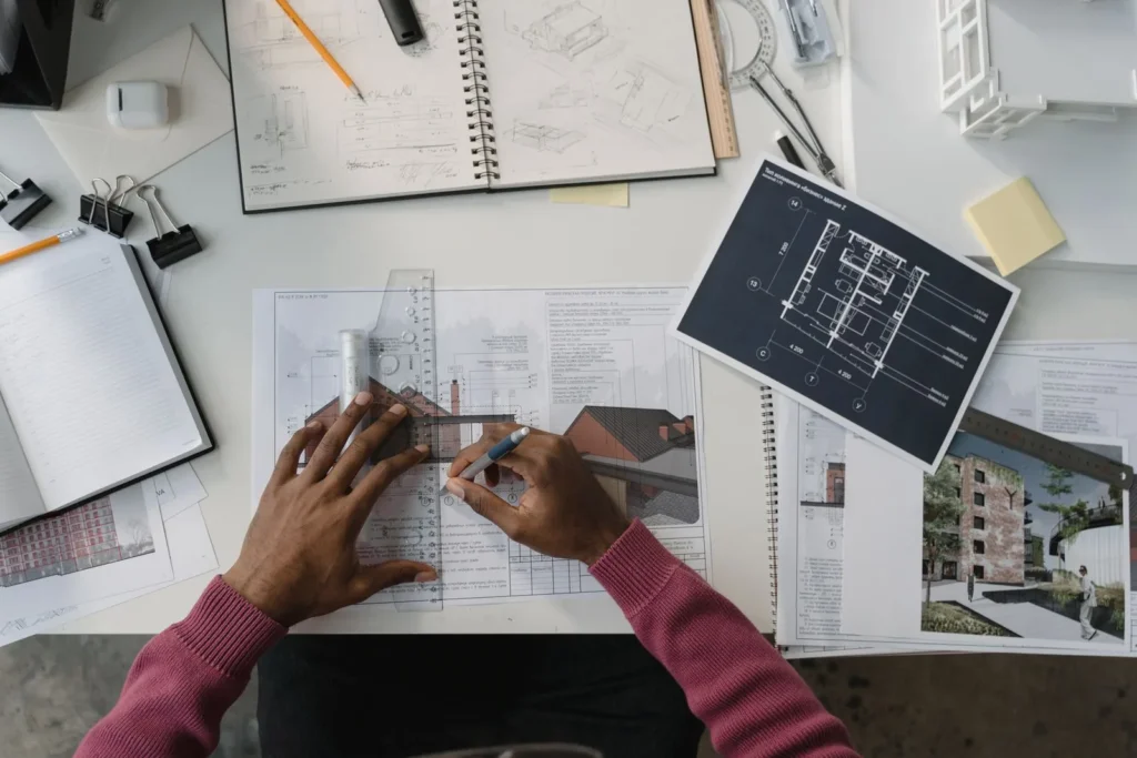 Hands drafting over printed drawings and renderings of a brick housing project spread across a desk