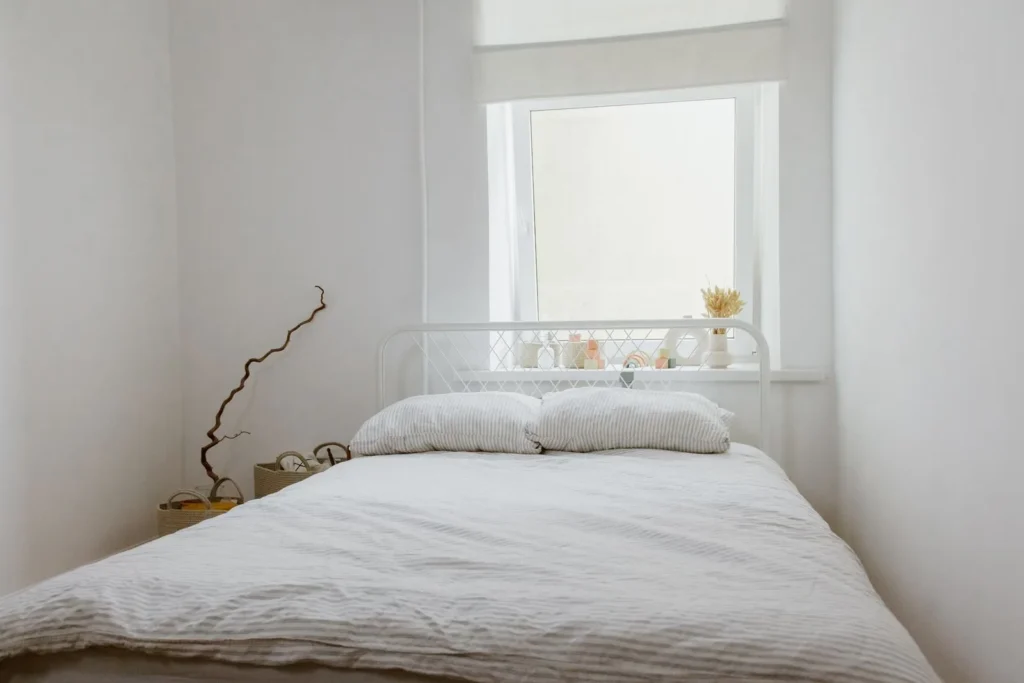 Simple white bedroom with a metal bed, linen bedding and a few dried branches by a sunny window