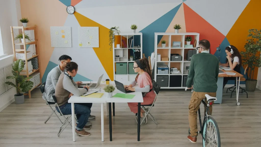 People working at shared tables in a bright coworking room with shelving and a colourful wall