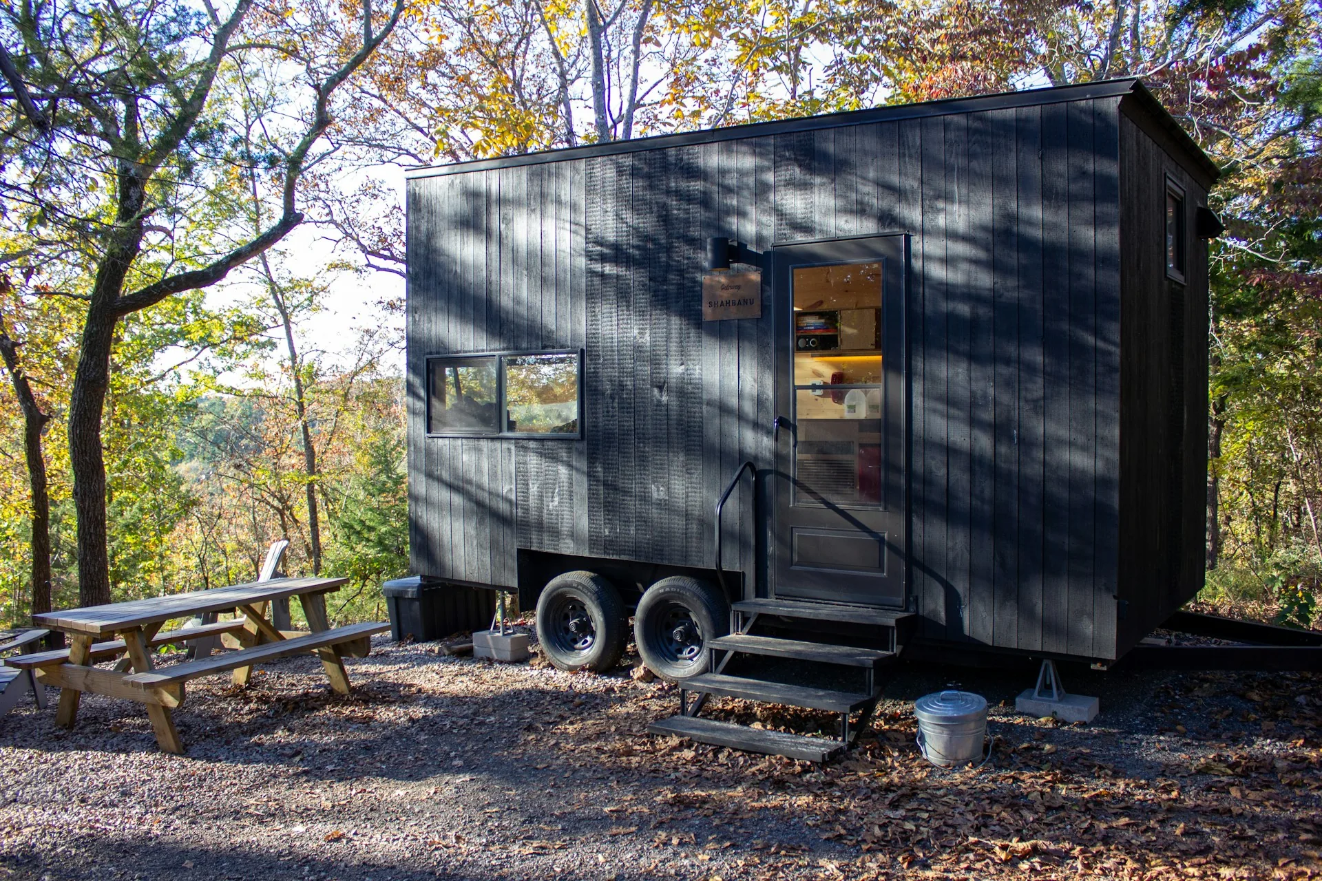 Dark timber tiny house on a dual-axle trailer parked at a wooded site, with a picnic table and entry steps