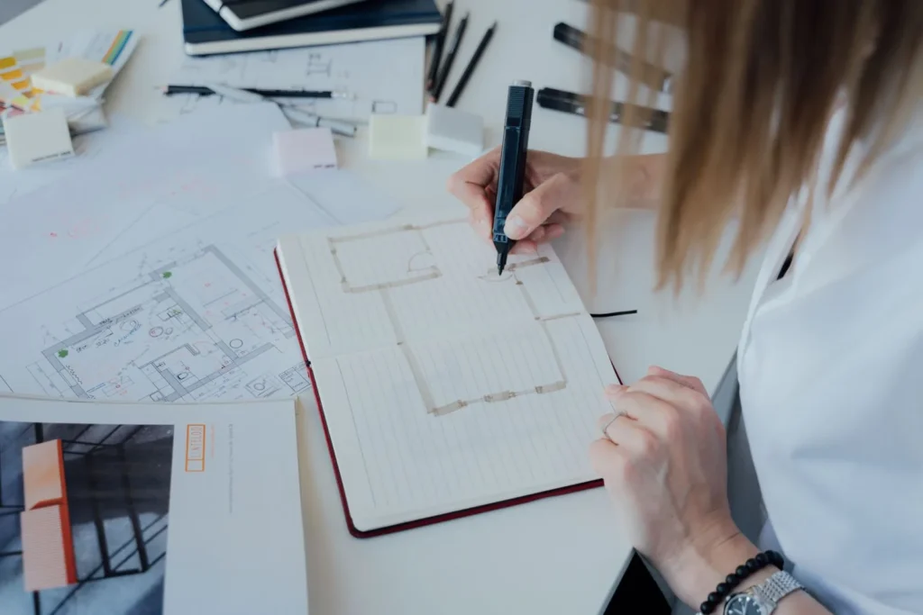 Design student sketching a room layout in a notebook surrounded by printed floor plans, a daily habit for interior architecture students