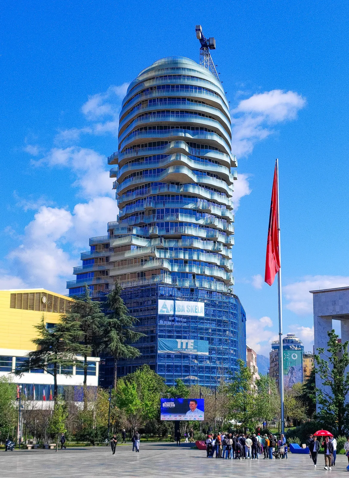 A curving residential tower with stacked rounded floors and planted terraces under a blue sky, with people in the square in front