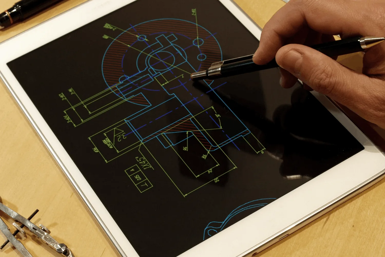 Pen tracing a coloured technical CAD drawing on a dark tablet screen beside drafting compasses