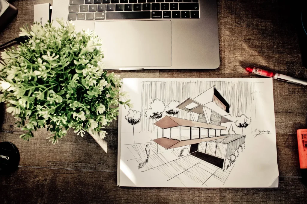 Hand-drawn perspective sketch of a modern house on a wooden desk beside a laptop and a plant