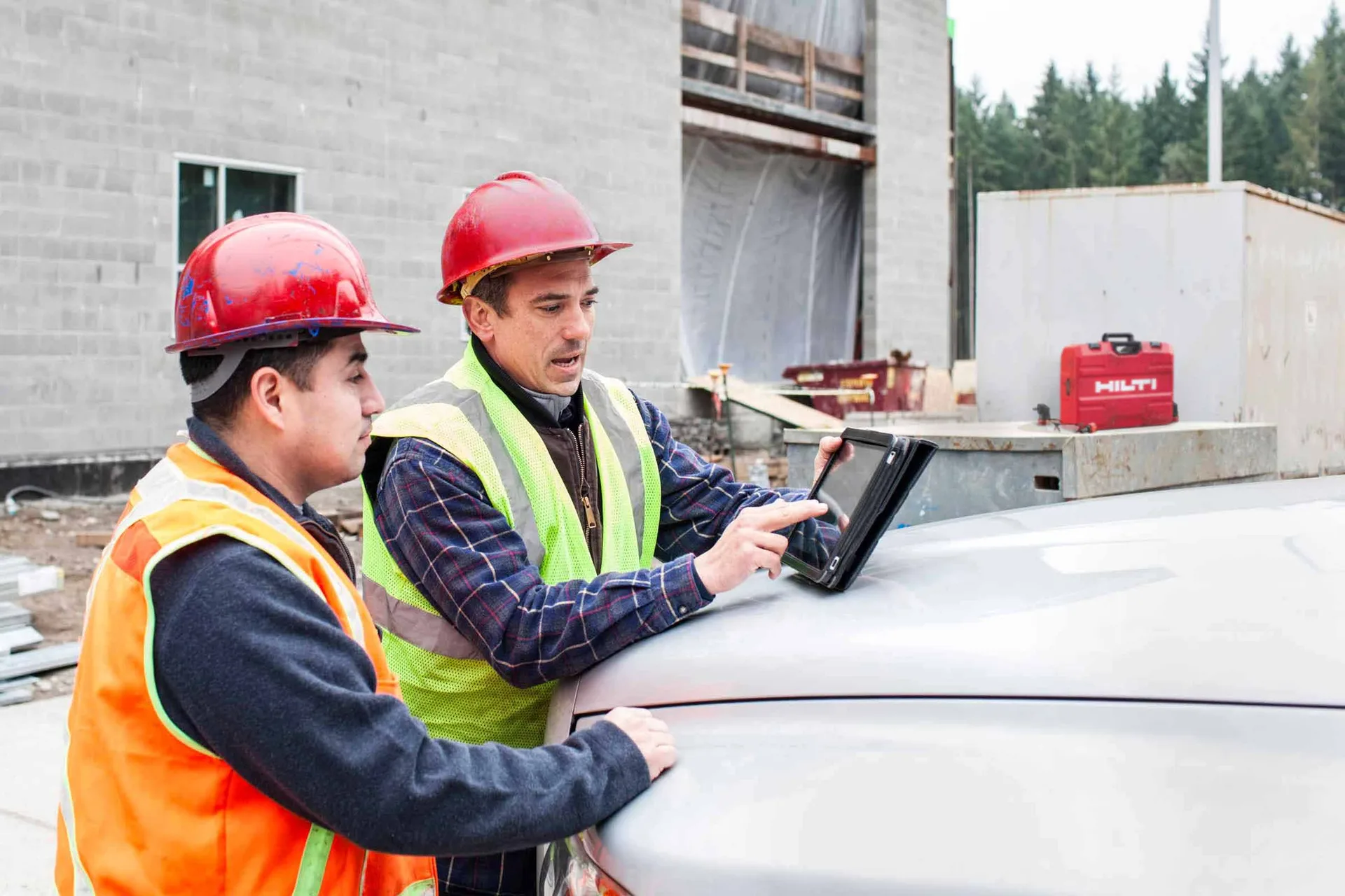 Two site workers in hard hats checking Fieldwire on a tablet beside a Hilti toolbox