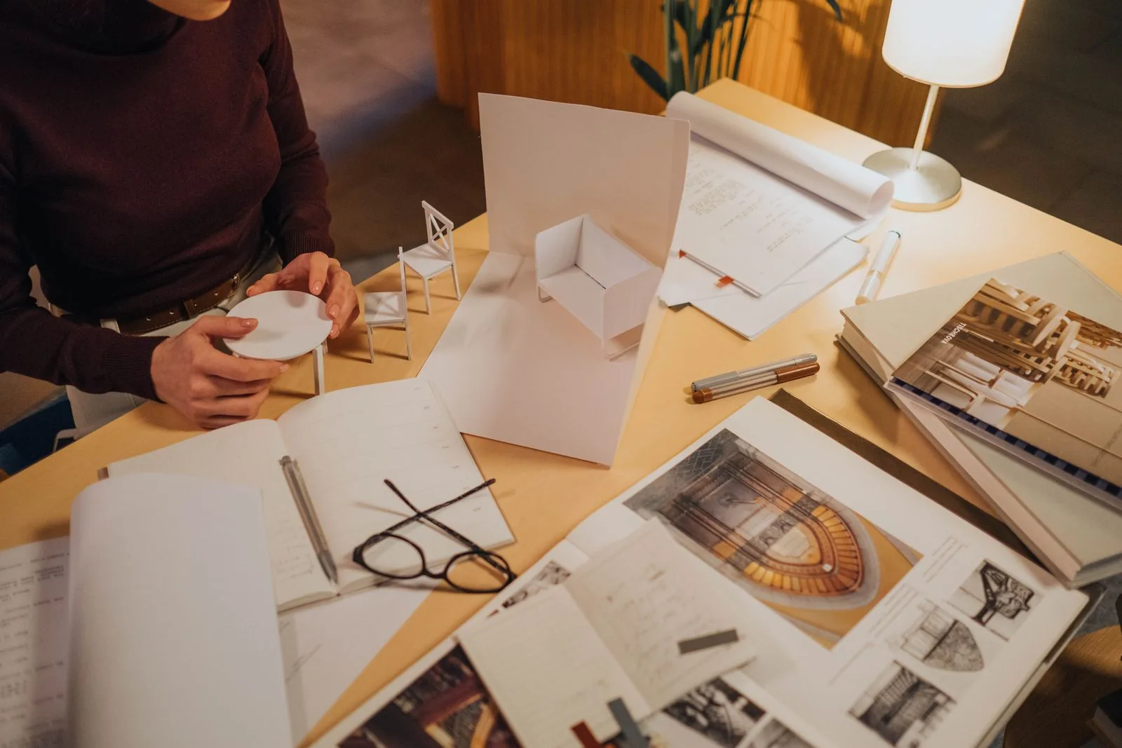 Hands assembling a white paper study model on a desk covered with sketches, drawings and reference books