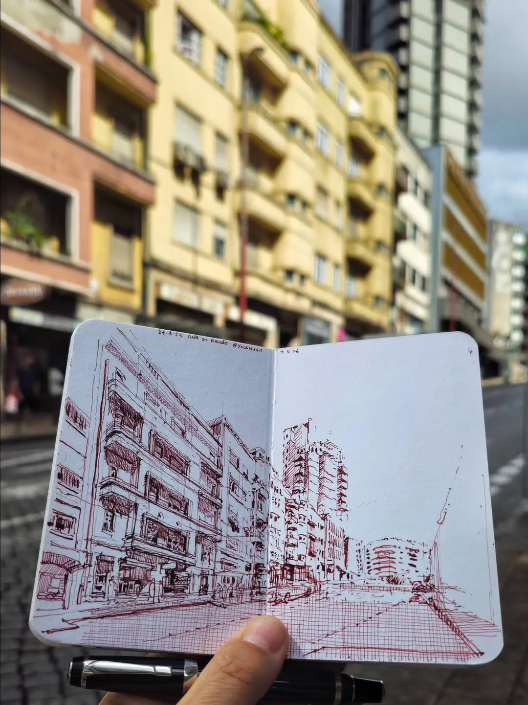 Sketchbook held up against the building it records, with a quick red ink facade study made on site
