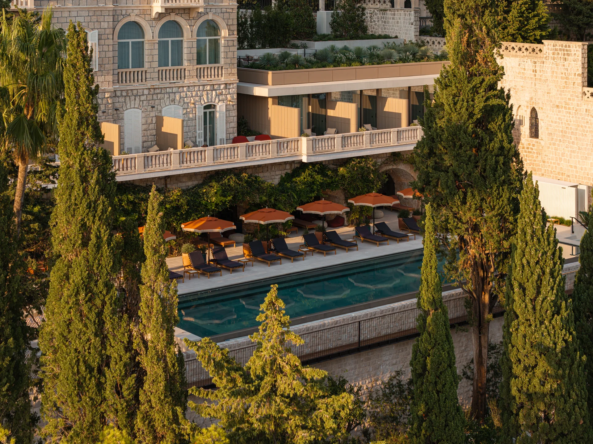 Historic stone villa with arched windows above an outdoor pool lined with orange parasols and tall cypress trees, Villa Argentina by 3LHD