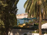 Terrace with green parasols between palm and pine trunks, looking across the water to the walls of Dubrovnik Old Town, Villa Argentina by 3LHD
