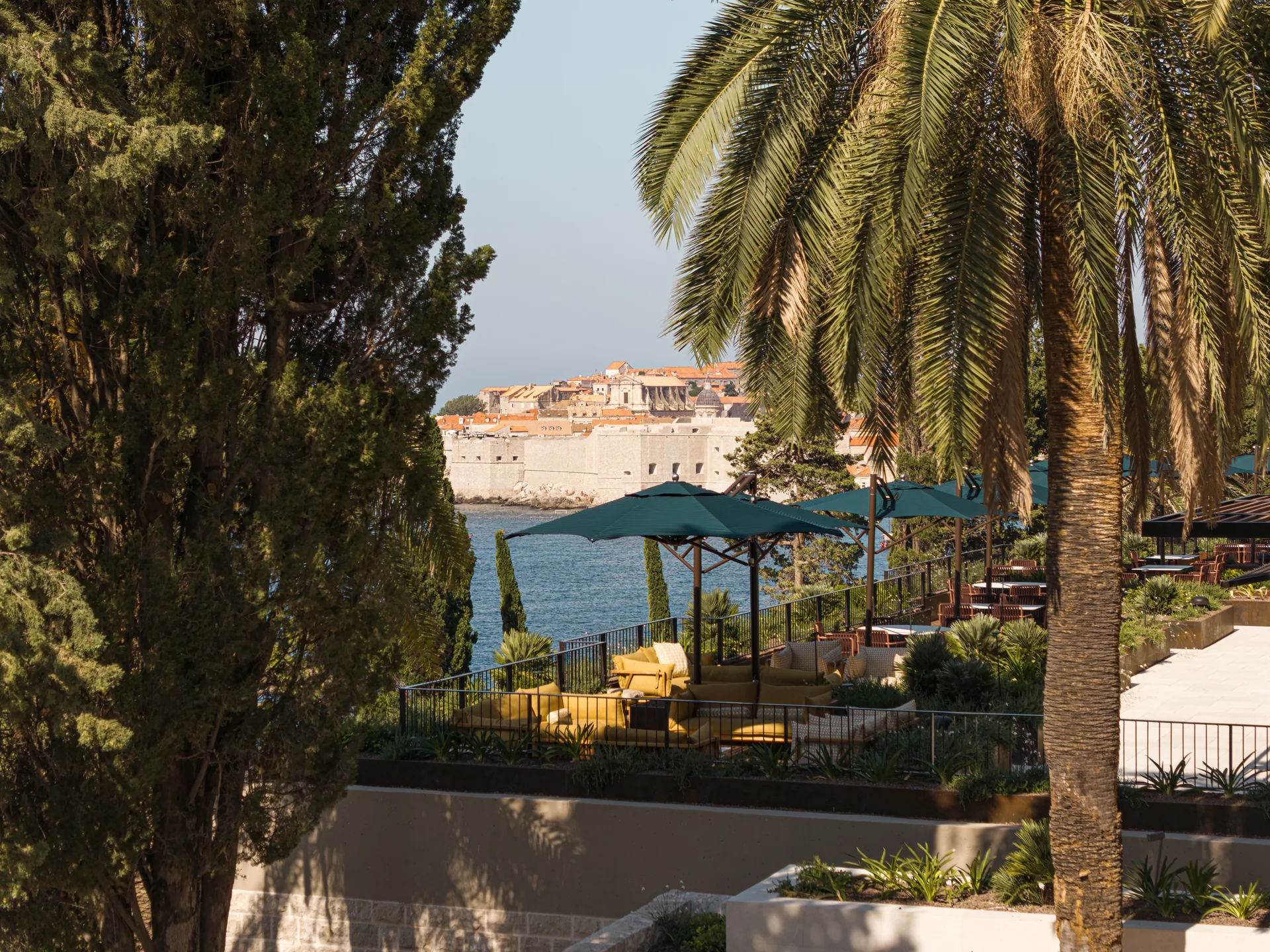Terrace with green parasols between palm and pine trunks, looking across the water to the walls of Dubrovnik Old Town, Villa Argentina by 3LHD