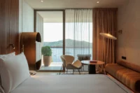 Guest room with a curved headboard, lounge chairs and a full height window opening to a balcony and the sea, Villa Argentina by 3LHD