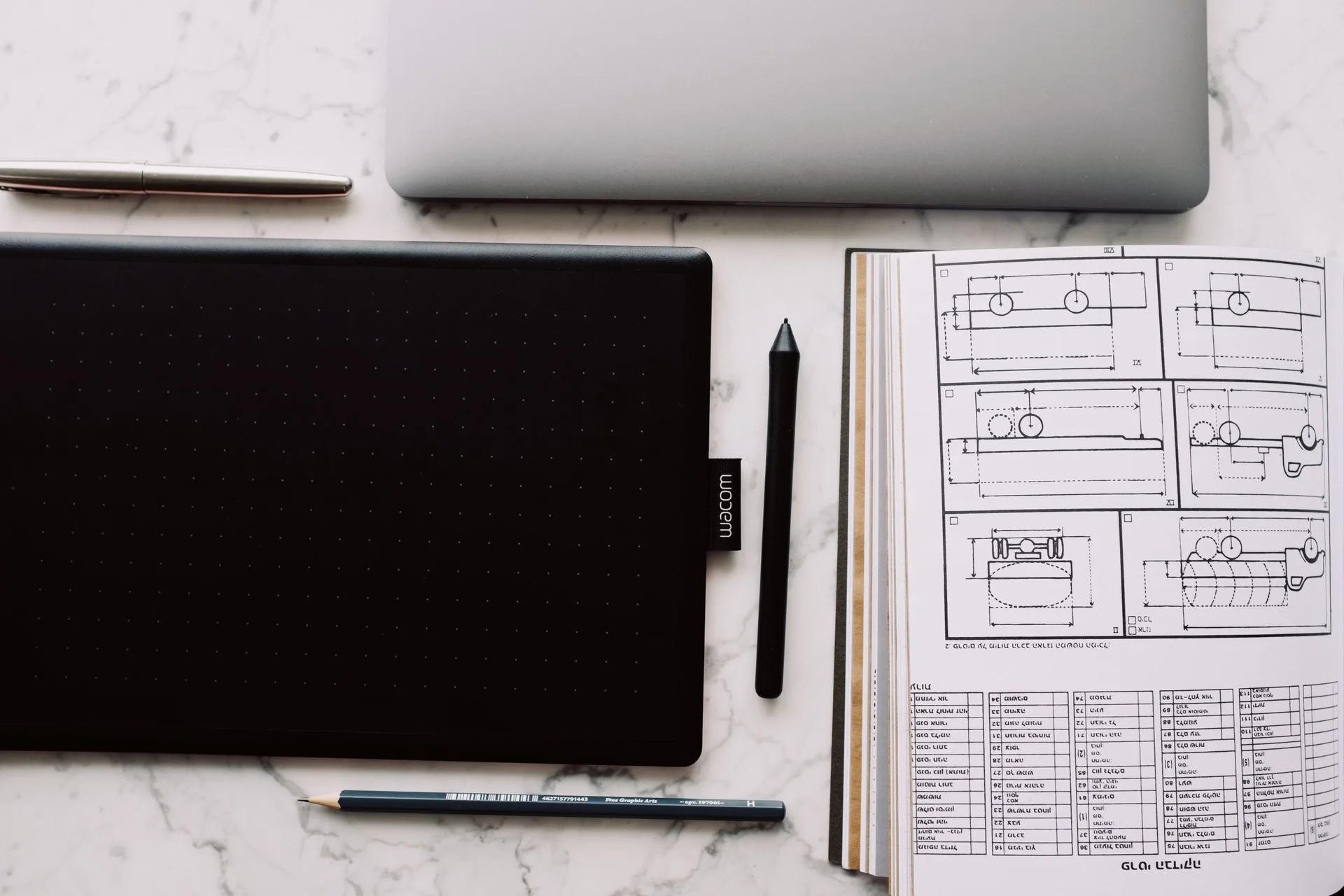 Wacom pen tablet and stylus beside an open sketchbook of technical drawings, a pencil and a fountain pen